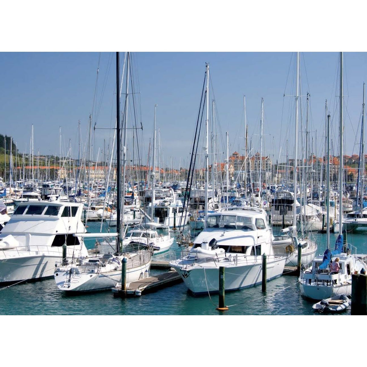 The marina is crowded with various vessels on a sunny day as a yacht using "Manoeuvring" by Adlard Coles glides effortlessly across calm waters, with buildings gleaming on the hillside against a clear blue sky.