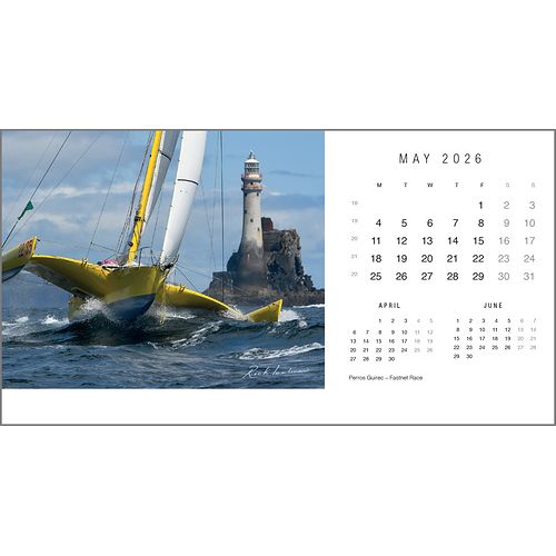 A yellow sailboat speeds past ocean waves and a rocky island with a lighthouse. On the right, the Rick Tomlinson Desk Calendar 2026 from Rick Tomlinson Photography displays May, reflecting his dynamic yachting style.