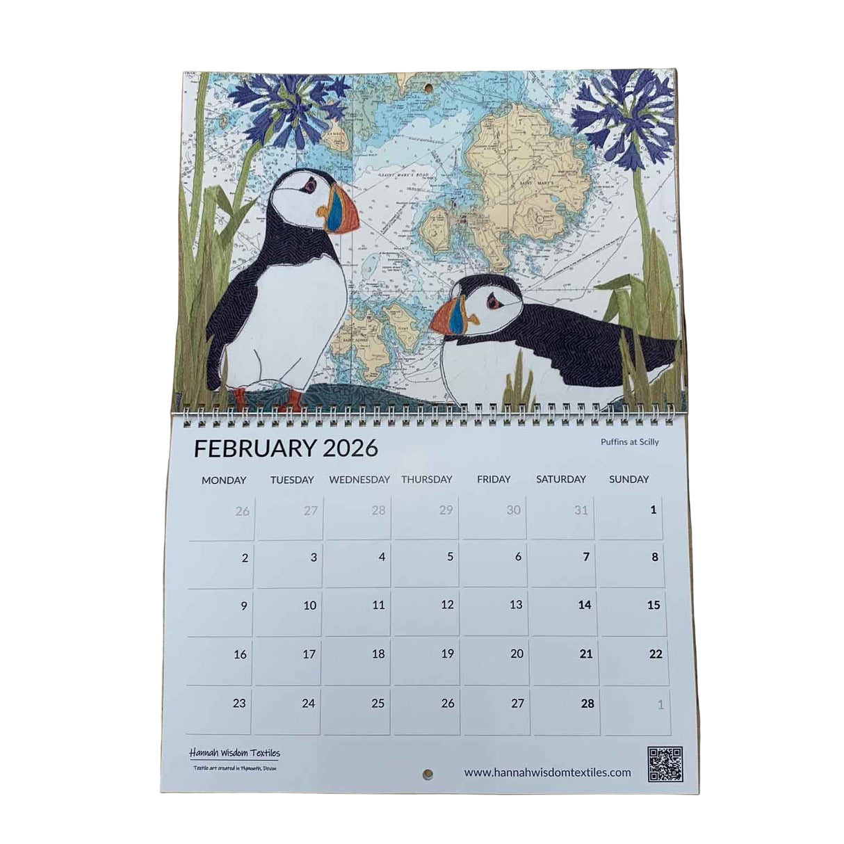 The Hannah Wisdom Coastal Calendar 2026 by Hannah Wisdom Textiles is open to February, displaying two puffins with flowers and a map-inspired, textile art background. The grid shows dates, days, and www.hannahwisdomtextiles.com at the bottom.