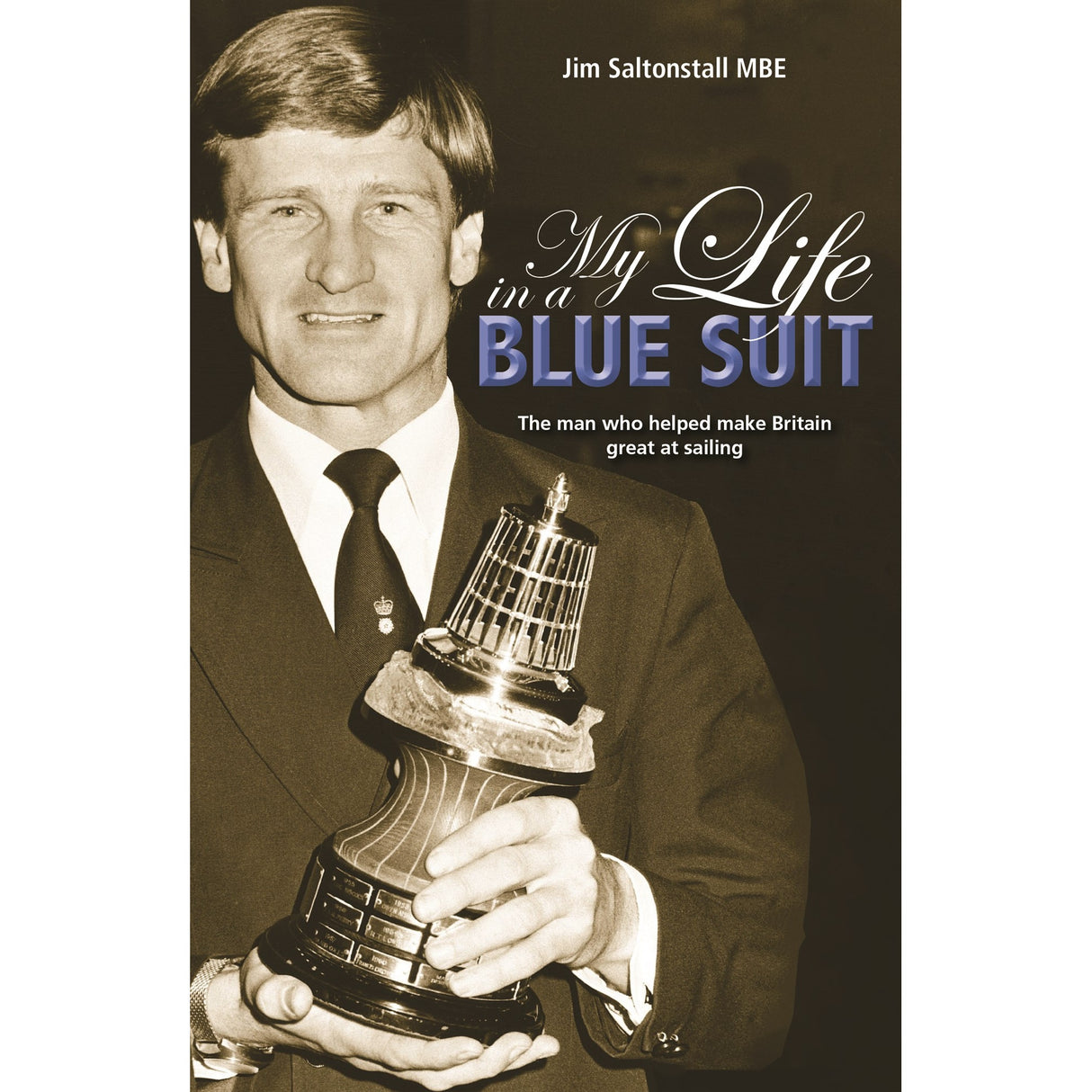 A man in a suit holds a large sailing trophy. The book cover reads: "My Life In A Blue Suit" by Fernhurst Books, telling the story of Olympic sailing coach Jim Saltonstall MBE, who helped make Britain great at sailing.