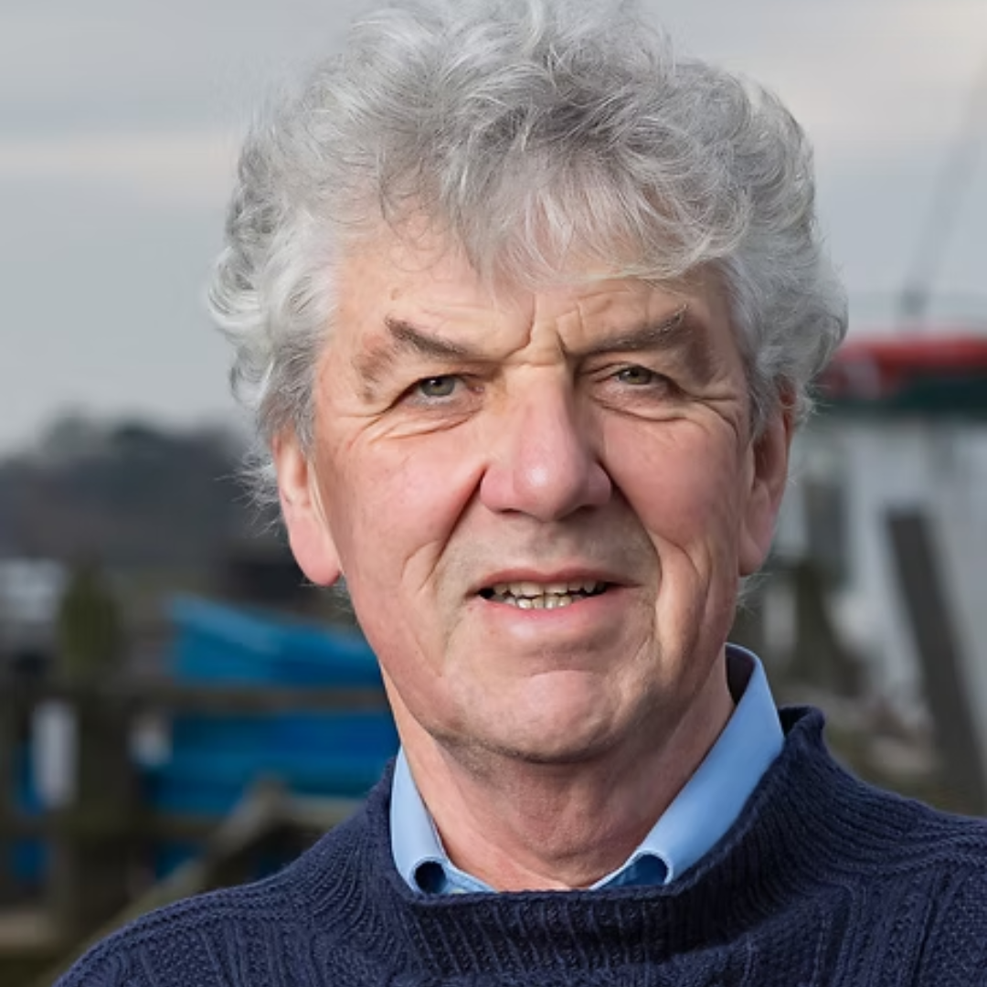 Portrait of an older man with gray curly hair, wearing a blue collared shirt and a navy sweater. He is outdoors, with a blurred background that suggests a harbor setting.