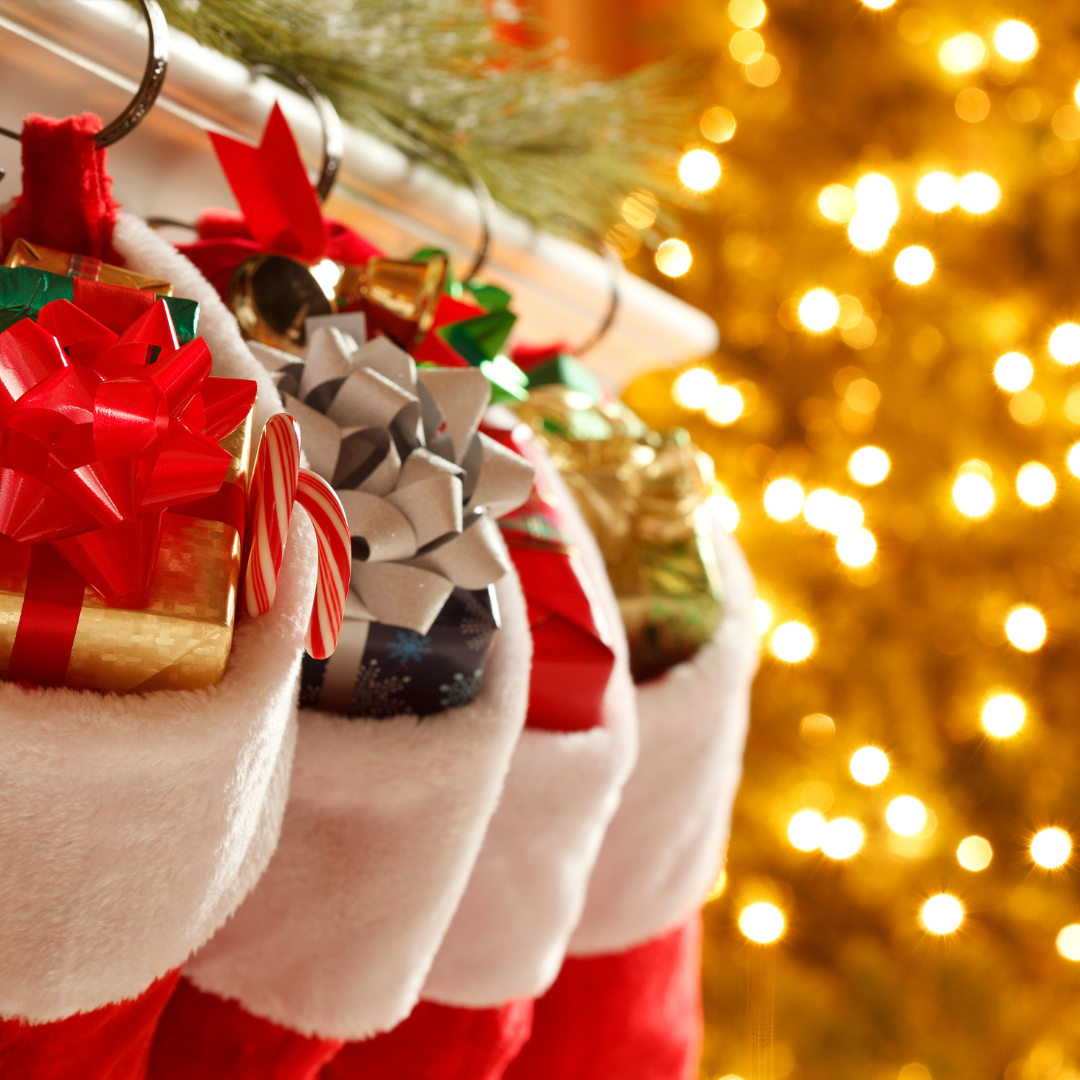 A row of Christmas stockings hanging on a mantelpiece, filled with presents There are out of focus fairy lights behind.