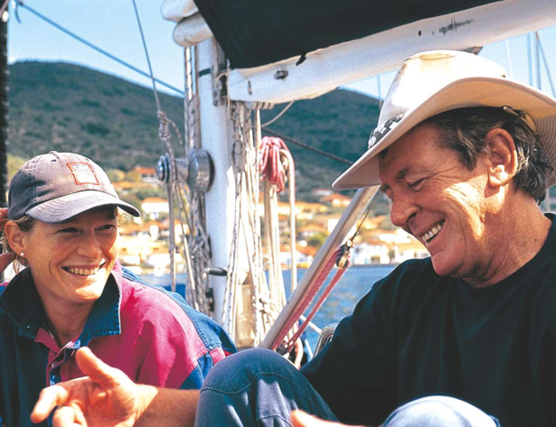 Two people smiling on a sailboat. The person on the left is wearing a baseball cap and jacket, while the person on the right is wearing a cowboy hat and black shirt. Hills and buildings are visible in the background.