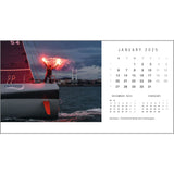 A sailor on a sailboat ignites a flare as the sun sets, highlighting the calm sea and distant shoreline. The Rick Tomlinson Desk Calendar 2025 by Rick Tomlinson Photography is on the right, with highlighted dates. The caption reads, "Dale Brauer – First American female solo circumnavigator.