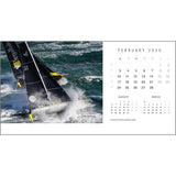 An exhilarating aerial shot showcases a sailing yacht braving turbulent ocean waves. To the right, the Rick Tomlinson Desk Calendar 2025 prominently displays dates and days for February, with smaller sections showing January and March. The scene highlights the remarkable skill of Rick Tomlinson Photography in capturing yachting adventures.