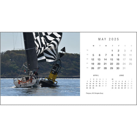 The Rick Tomlinson Desk Calendar 2025 by Rick Tomlinson Photography features a stunning image for May 2025, showcasing a sailboat with black and white sails gliding near a yellow buoy on a tranquil sea. The scene, expertly captured by renowned yachting photographer Rick Tomlinson, includes a forested coastline in the background and offers views of the previous and next months.