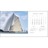 A sailboat with white sails glides across the water under a blue sky. On the right, a Rick Tomlinson Desk Calendar 2025 from Rick Tomlinson Photography is displayed, highlighting weekends in bold and evoking the yachting captures by Rick Tomlinson.