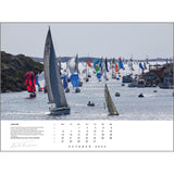 A vibrant sailing race with colorful sails unfurled, captured in stunning sailing photography by Rick Tomlinson Photography. Rocky shorelines and houses form the backdrop, while a page from the Rick Tomlinson Portfolio Calendar 2025 for October highlights this picturesque scene on water.