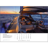 A sleek yacht glides on the ocean at sunset, warm sunlight illuminating its deck and controls. The sky is a mix of orange and blue hues. This captivating scene adorns the December 2025 page of the Rick Tomlinson Portfolio Calendar 2025 by Rick Tomlinson Photography, featuring exquisite sailing photography.