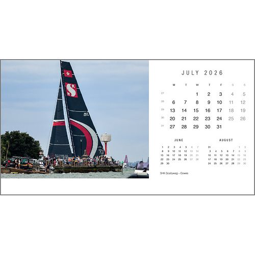 A large sailboat with a black and red sail glides past a busy pier under a clear sky in Rick Tomlinson's iconic yachting photo. On the right is the July 2026 Rick Tomlinson Desk Calendar by Rick Tomlinson Photography, with June and August below.