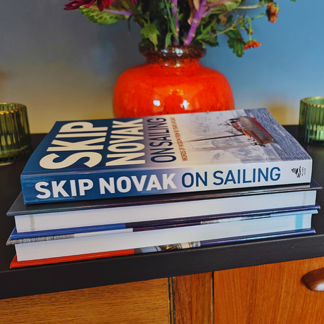 A stack of books rests on a wooden sideboard, adorned with a vibrant orange vase and flowers in the background. The top book, "Skip Novak on Sailing" by Adlard Coles, provides insights into expert seamanship. Flanking the scene are two green glass candles, capturing a serene moment reminiscent of Yachting World.