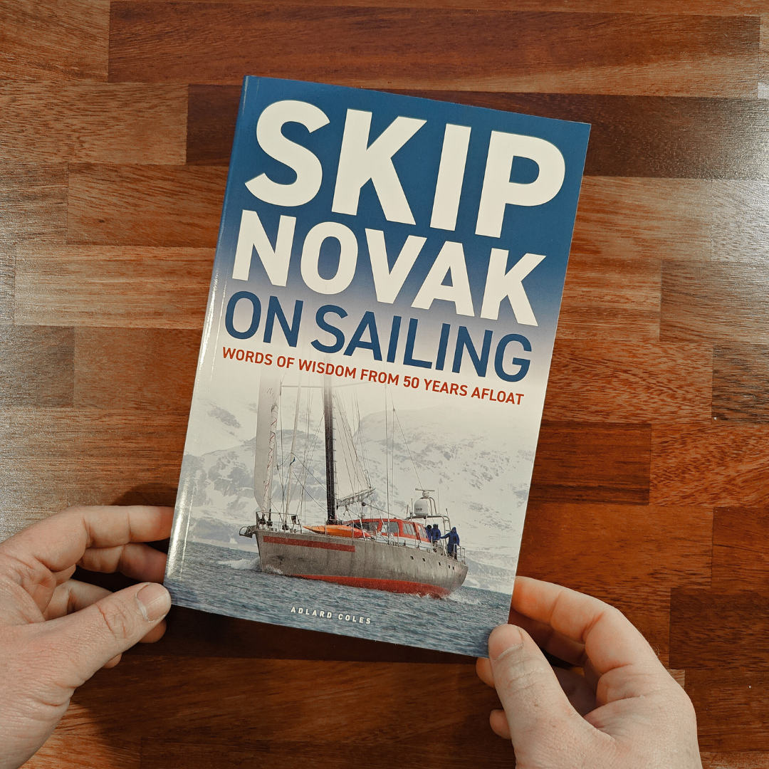 Two hands hold the Adlard Coles book, "Skip Novak on Sailing," which features an image of a sailboat on the cover. The wooden background suggests the rich seamanship and celestial navigation stories contained within its pages.