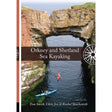 Two people kayak in clear blue water under a rock arch with cliffs and green grass. The book cover shows "Orkney and Shetland Sea Kayaking" by Pesda Press, with the authors’ names listed at the bottom.