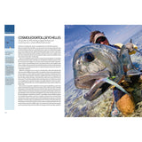 A fly fisher in sunglasses and waders stands in clear water behind a large fish hooked on a lure—a perfect moment from Ultimate Fishing Adventures by Fernhurst Books, captured under a blue sky with scattered clouds.