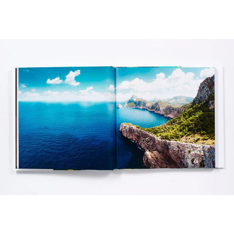 An open book, "The Life and Love of the Sea" by Abrams Books, showcases a stunning aerial photograph of a rugged coastline with lush green hills and deep blue ocean beneath a bright sky dotted with clouds, capturing the vivid beauty of marine landscapes and the enchanting world depicted in underwater photography.