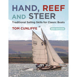 A classic gaff rig sailboat glides on water against hilly terrain under a cloudy sky. Text reads: "Hand, Reef and Steer," by Adlard Coles, 2nd Edition, traditional sailing skills for classic boats.