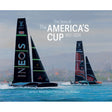 Two racing sailboats with large sails glide across the water, cityscape in the background. Text reads: “The Story of The America’s Cup 1851–2024.” This captivating book from ACC Art Books, featuring artwork by Tim Thompson and written by Ranulf Rayner, beautifully captures the rich sailing history of this legendary yacht race.