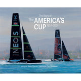 Two racing sailboats with large sails glide across the water, cityscape in the background. Text reads: “The Story of The America’s Cup 1851–2024.” This captivating book from ACC Art Books, featuring artwork by Tim Thompson and written by Ranulf Rayner, beautifully captures the rich sailing history of this legendary yacht race.