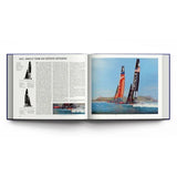 An open copy of "The Story of the America’s Cup 1851-2024" by ACC Art Books displays a page featuring text about Oracle Team USA alongside a painting of two racing sailboats on the water, set against a backdrop of a city and bridge—capturing an iconic moment from America’s Cup sailing history.