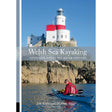 A kayaker paddles along a rocky shoreline beneath a white and red lighthouse under a clear blue sky. Text reads: "Welsh Sea Kayaking" by Pesda Press—your essential guide to 51 great sea kayak voyages for exploring the stunning Welsh coastline.