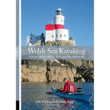 A kayaker paddles along a rocky shoreline beneath a white and red lighthouse under a clear blue sky. Text reads: "Welsh Sea Kayaking" by Pesda Press—your essential guide to 51 great sea kayak voyages for exploring the stunning Welsh coastline.