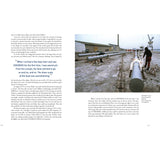 In an industrial dock dusted with snow, two people work on a long sailboat mast amid warehouses and cranes—an image reminiscent of Dick Carter: Yacht Designer by Fernhurst Books.