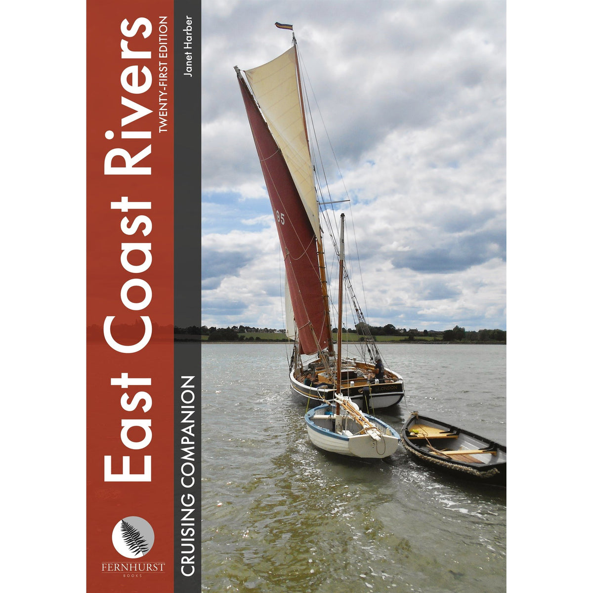 A sailboat with a large reddish-brown mainsail towing two small wooden dinghies on a river, as featured in the East Coast Rivers Cruising Companion by Fernhurst Books.