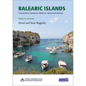 Cover of the Imray "Balearic Islands" sailing pilot book features sailboats anchored in clear blue water between rocky cliffs with greenery and a partly cloudy sky. Title and authors' names appear at the top.