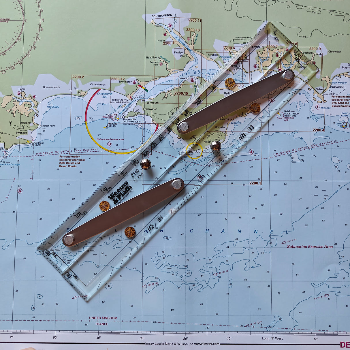 A navigation chart displaying coastal and nautical details with a durable Weems & Plath 12" Parallel Rule placed on it. The rule, featuring metal screws and an engraved protractor scale, is used to mark courses on the map.