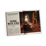 An open magazine features an article titled "Fire to Fork," published by Exploring Eden Media. On the right, someone is honing their campfire cooking skills amid sand and rocks at night. The left page hints at outdoor cuisine with captivating text introducing the piece.