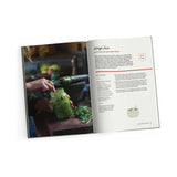 An open copy of "The Small Kitchen Cook" from Exploring Eden Media features a person using a spoon to scoop vibrant pesto from a jar for the "Raw Green Pesto" recipe. Fresh herbs and a mortar and pestle decorate the table, making it perfect for individuals with compact kitchens. On the right page, the detailed recipe provides creative cooking techniques.