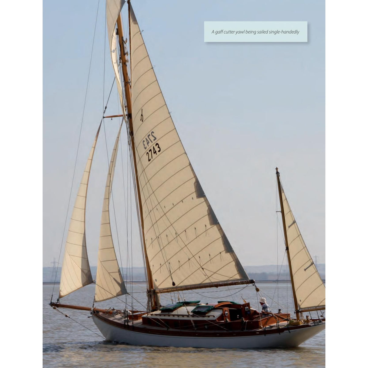 A gaff cutter yawl by Adlard Coles called Stress-Free Sailing glides on calm waters with cream sails and a wooden deck. A person is visible at the helm, mastering the steady course under a light blue sky.