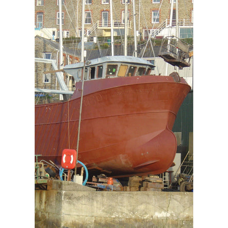 A spacious red powerboat, featuring the advanced hull design from Adlard Coles' "Powerboat Design and Performance," is docked on a dry platform in a shipyard. The background displays multi-story brick buildings alongside several other boats, with a life preserver visible in the foreground.