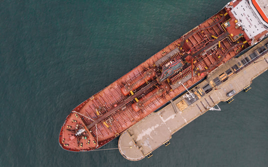 Aerial view of a large red cargo ship docked at a pier. The ship is loaded with various containers and cargo secured with ropes and equipment. The surrounding water is calm and deep green in color. The pier has multiple platforms extending out into the water.