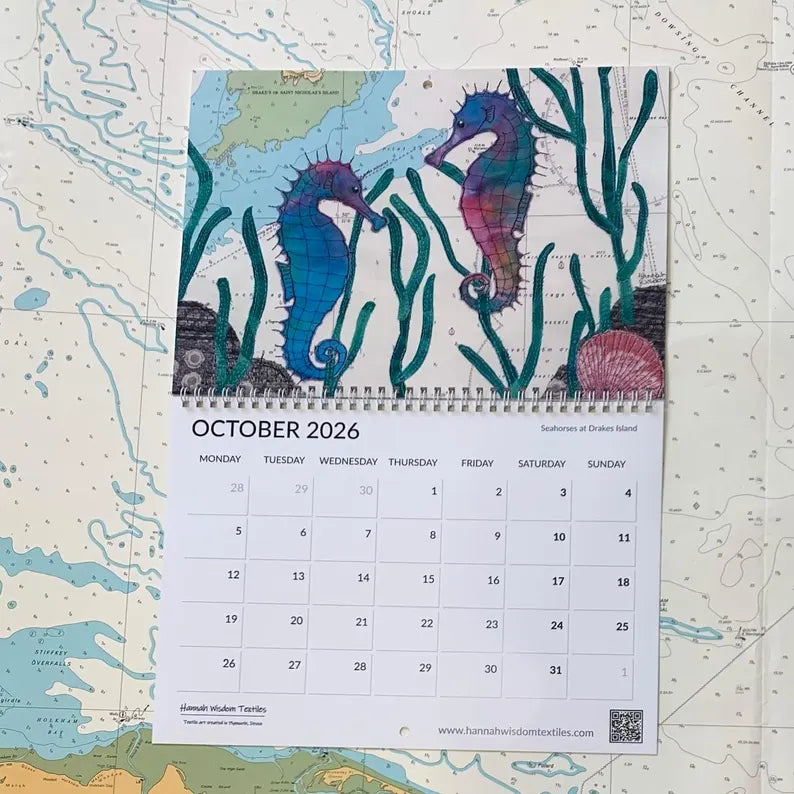 The Hannah Wisdom Coastal Calendar 2026 by Hannah Wisdom Textiles hangs open to October, displaying colorful seahorses among seaweed at the top, with the month's days visible below the artwork.