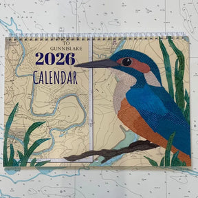 The "Hannah Wisdom Coastal Calendar 2026" by Hannah Wisdom Textiles features a kingfisher among green grass on a detailed map, reflecting the signature illustrated style of the brand.