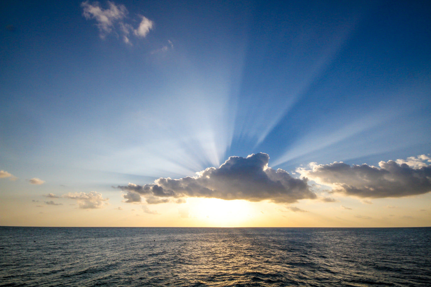 A serene ocean view at sunset with sun rays streaming from behind clouds, casting a warm glow on the water. The sky transitions from blue to shades of orange near the horizon.